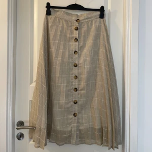 Midi Collection Beige rutig långkjol - Snygg beige långkjol med vitt rutmönster från Mudo Collection. Kjolen har knappar framtill och är gjord i ett luftigt linnematerial, perfekt för varma dagar. Den har en avslappnad passform och ger en chill vibe till din outfit.