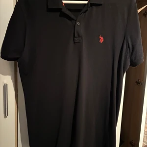 Svart pikétröja från Ralph Lauren - Klassisk svart pikétröja från Ralph Lauren med röd broderad logga på bröstet. Tröjan har korta ärmar, krage och knappar vid halsen. Perfekt för en clean och stilren look.