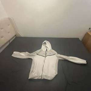 Vit hoodie från Nike med blå logga - Säljer en vit hoodie från Nike i storlek M. Tröjan har hel dragkedja, huva och svarta detaljer längs bröstet. Nike-loggan är broderad i blått på vänster sida. Perfekt för en sportig och avslappnad stil.