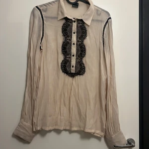 Beige skjorta med spets från Gina Tricot - Säljer en beige skjorta från Gina Tricot med svarta spetsdetaljer längs knappraden och axlarna. Skjortan har klassisk krage, långa ärmar och svarta knappar framtill. Tunt och lätt material som ger en luftig känsla.