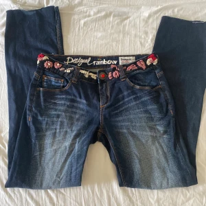 Desigual jeans med broderade blommor - Mörkblå jeans från Desigual med broderade blommor i rosa, rött och vitt längs linningen. Klassisk femficksmodell med kontrastsömmar och en unik knapp med röd detalj. Regular fit och snygg tvättad effekt framtill och baktill. Strl 30, Fraktar gärna tillsammans med andra plagg.