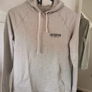Grå hoodie från Lexington - Snygg grå hoodie från Lexington med vit snörning och tryck på bröstet. Tröjan har huva, lång ärm och en klassisk känguruficka framtill. Perfekt för chill dagar och enkel att matcha med jeans eller mjukisbyxor.