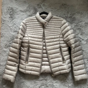 Beige pufferjacka med dragkedja - Säljer en beige pufferjacka med horisontella quiltade partier och hög krage. Jackan har två fickor med dragkedja och en stilren, minimalistisk look. Perfekt för kyliga dagar och enkel att matcha med olika outfits. Size M💕