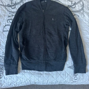 Mörkgrå kofta från Polo Ralph Lauren - Snygg mörkgrå kofta från Polo Ralph Lauren med dragkedja framtill och broderad logga på bröstet. Tröjan har långa ärmar, ribbade muddar och två fickor med dragkedja. Perfekt för dig som gillar stilrena och klassiska plagg.