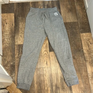 Grå mjukisbyxor med snörning - Säljer ett par grå mjukisbyxor med resår och snörning i midjan. Byxorna har en avslappnad passform, muddar vid bensluten och en liten vit logga på vänster sida. Perfekta för chill dagar eller när du vill vara bekväm.