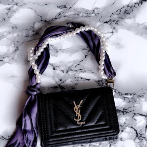 Svart YSL handväska med pärlhandtag - Snygg svart quiltad handväska från YSL med guldigt logospänne. Handtaget är dekorerat med lila scarf och pärlor för en unik och trendig look. Perfekt accessoar för att lyfta din outfit. Inte äkta men jättebra qualitet. Mini väska 