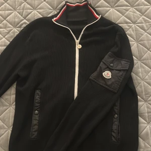 Svart Moncler kofta med dragkedja - Svart ribbad kofta från Moncler med hel dragkedja och hög krage med röd och vit rand. Fickor med knappar och en snygg Moncler-logga på ärmen. Perfekt för dig som gillar stilrena och exklusiva plagg. Storleks lappen har försvunnit men det är storlek L