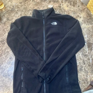 Svart fleecejacka från The North Face - Svart fleecejacka från The North Face med hel dragkedja. Perfekt för kyliga dagar och enkel att matcha med andra plagg. Storlek S herr. Köpt för 1200kr. Hör av er vi fler frågor!