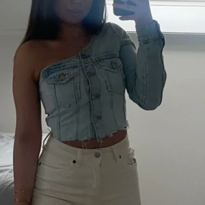 Croppad jeansbolero med oneshoulder - Säljer en ljusblå croppad bolerotopp i jeansmaterial med oneshoulder-design och lång ärm. Toppen har knappar framtill, rå kant nedtill och coola sömmar samt fickdetaljer på bröstet. Perfekt för dig som gillar unik stil och denim.