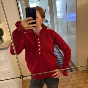 Röd stickad tröja med pärlknappar - Mysig röd stickad tröja med volangkant längs halsringningen och pärlknappar framtill. Tröjan har långa ärmar med ribbade muddar och en feminin touch tack vare de dekorativa detaljerna. Perfekt för dig som gillar en söt och trendig stil.