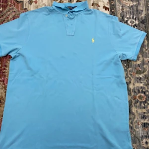 Pikétröja från Polo Ralph Lauren - Snygg ljusblå pikétröja från Polo Ralph Lauren med klassisk krage och två knappar framtill. Ikoniska gula loggan broderad på bröstet. Tillverkad i bomull för en skön och luftig känsla.