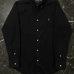 Svart skjorta från Polo Ralph Lauren - Svart Polo Ralph Lauren skjorta. Button-down, röd logga. Storlek S/M, sitter mer som S. Den fixar allt, från möte/skola till helgkväll. Det måste verkligen lägga till den i garderoben. Skicket är som nytt. Inget att klaga på. Köp den nu!