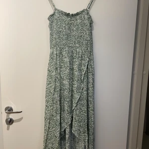 Grön blommig maxiklänning från SHEIN - Lång grön klänning från SHEIN med smala axelband och blommigt mönster. Smockad överdel och flowy kjol med omlottkänsla och volangdetalj framtill. Perfekt för varma dagar och somriga vibes.