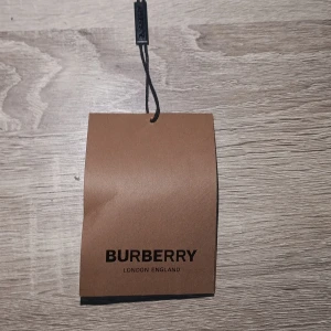 Burberry väska med klassisk tag - Säljer en exklusiv väska från Burberry, London England. Väskan har en klassisk brun tag med svart text och elegant snöre. Perfekt för dig som gillar lyxiga accessoarer och tidlös design.