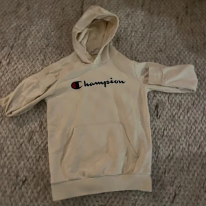 Beige hoodie från Champion, storlek L - Säljer en beige hoodie från Champion i storlek L. Tröjan har klassisk huva, magficka och stor logga framtill. Perfekt för dig som gillar streetwear och vill ha en enkel men snygg look.