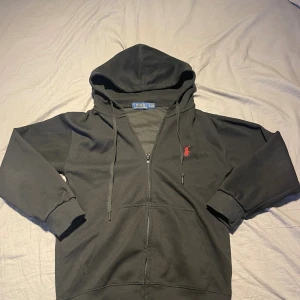 Svart hoodie från Polo Ralph Lauren - Svart hoodie från Polo Ralph Lauren med dragkedja och huva. Klassisk röd broderad logga på bröstet, ribbade muddar och fickor framtill. Tillverkad i mjuk bomullsmix, perfekt för chill dagar eller när du vill ha en clean streetstyle.