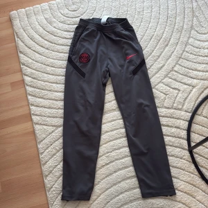 Nike PSG Dri-Fit träningsbyxor grå - Grå träningsbyxor från Nike med PSG-logga och röd swoosh på framsidan. Byxorna är tillverkade i Dri-Fit polyester, har elastisk midja och raka ben. Perfekta för fotboll eller chill, med snygga svarta detaljer vid fickorna.