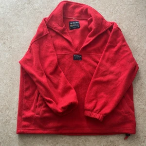 Röd fleece-tröja från Norman Wells - Riktigt skön röd fleece-tröja från Norman Wells med hög krage och långärmade ärmar. Tröjan har en enkel design med elastiska muddar och dragsko nedtill. Perfekt för kyliga dagar och enkel att slänga på sig när man vill vara varm.