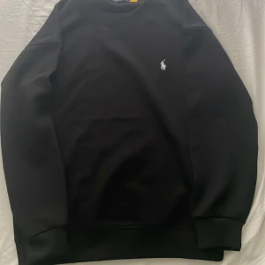 Svart sweatshirt från Polo Ralph Lauren - Svart sweatshirt från Polo Ralph Lauren med klassisk rund hals och broderad vit logga på bröstet. Tröjan har långa ärmar och ribbade muddar vid hals, ärmslut och nederkant. Perfekt för en clean och stilren look. Det är storlek M men passar mer som S eller även xs.