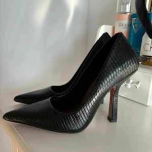 Svarta pumps med spetsig tå - Säljer ett par svarta pumps med hög klack och spetsig tå. Skorna har ett stilrent, diskret mönster i materialet som ger en lyxig känsla. Tillverkade i skinnimitation med en elegant finish, perfekta för dig som vill sticka ut med klassiska men trendiga klackar.