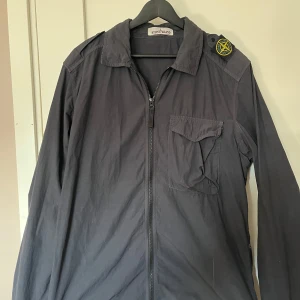 Stone Island Svart/Blå tunn overshirt  - Svart stone island overshirt. Perfekt inför hösten och våren. Nypris 3099kr. Ser svart ut men enligt NK är den blå. Står storlek L men passar mer som M.