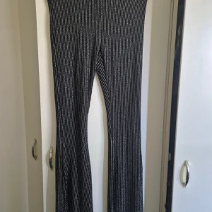 Svarta glittriga bootcut-byxor STAY M - Säljer ett par svarta bootcut-byxor från STAY i storlek M. Byxorna har smala vertikala silverglittriga ränder och är tillverkade i ett stretchigt syntetmaterial. De har hög midja och en riktigt snygg passform som ger en cool vibe.