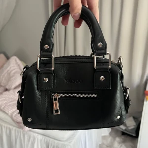 Svart handväska från Núnoo i skinn - Snygg svart handväska från Núnoo i skinn med silverfärgade metalldetaljer och kedjerem. Väskan har både handtag och axelrem, samt en dragkedja framtill och på toppen. Rymmer extremt mycket för att vara så liten. Nypris 1200kr.