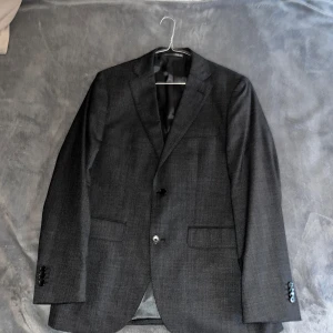 Mörkgrå kostym från Riley i ull - Säljer en stilren mörkgrå kostym från Riley i klassisk modell. Kavajen har två knappar, smala slag och innerfoder i svart. Byxorna är raka med pressveck och hällor för bälte. Perfekt för dig som vill ha en tidlös och snygg look. Enbart använt vid ETT tillfälle. Köpta från Brothers. Dessa har också ett nypris på: 3000kr. 