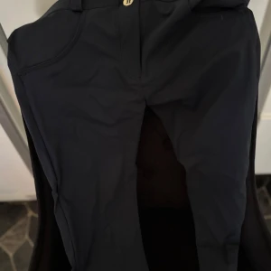 Svarta WR.UP byxor från Freddy, stl L - Svarta Freddy WR.UP byxor i storlek Large med snygg shaping-effekt och markerade bakfickor. Byxorna har hög midja, bälteshällor och är tillverkade i ett stretchigt syntetmaterial som sitter tight och formar kroppen.