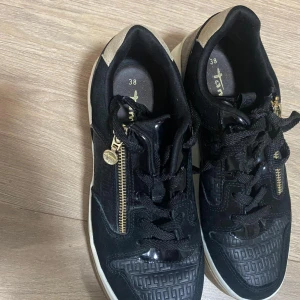 Svarta sneakers från Tamaris med guldzip - Snygga svarta sneakers från Tamaris med coola detaljer som guld dragkedja på sidan och mönstrade partier i svart och brunt. Skorna har snörning, platt vit sula och inslag av mocka och syntet. Perfekt för dig som gillar en sportig och trendig look.