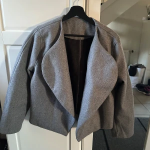 Grå kappa - Snygg grå kappa med omlottfront och mjukt, lite tjockare material. Kappan har lång ärm, rak passform och stilren design utan synliga knappar eller dragkedja. Strl S