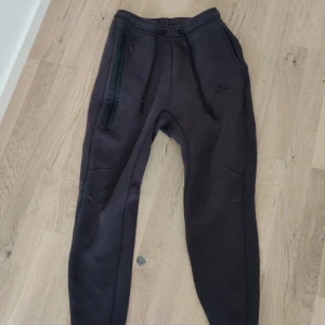 Nike tech byxor - Svarta mjukisbyxor från Nike med dragsko i midjan och diskret logga på benet. Byxorna har en smal passform, ribbade muddar vid bensluten och en dragkedjeficka på ena benet. Perfekta för chill eller träning.