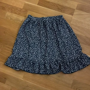 Mörkblå blommig volangkjol - Supersöt mörkblå kjol med vitt blommönster och volang längst ner. Resår i midjan för skön passform. Perfekt för dig som gillar en feminin och lekfull stil. Lätt och luftig, passar bra till sneakers eller sandaler.