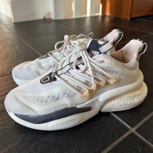 Adidas vita sneakers med boost-sula - Snygga vita Adidas sneakers med chunky boost-sula och svarta detaljer. Ovandelen är i mesh och syntet med coola linjer och snörning framtill. Bak finns en bred randig hälflik för enkel påtagning. Perfekta för dig som gillar sportig och modern stil.