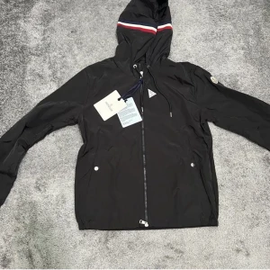 Svart vindjacka från Moncler med huva - Svart vindjacka från Moncler med klassisk logga på ärmen och huva med röd, vit och blå rand. Jackan har dragkedja framtill, två fickor med tryckknappar och justerbar dragsko i huvan. Tillverkad i lätt och smidig polyester, perfekt för blåsiga dagar.