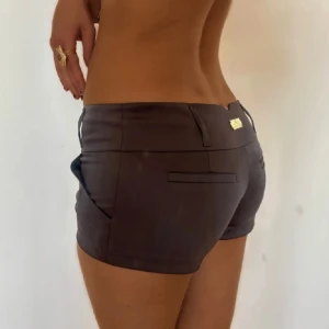 lågmidjade shorts  - Säljer ett par bruna lågmidjade shorts med snyggt satin. De har bälteshällor, dekorativa bakfickor och en liten metallbricka med logga bak. Midjemåttet är 45cm