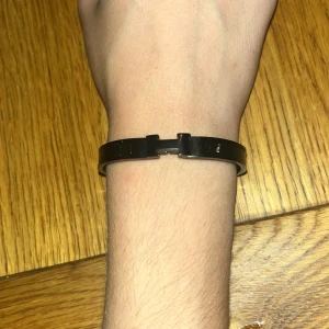 Svart armband med H-detalj - Stilrent svart armband i metall med en snygg H-formad detalj framtill. Armbandet har en enkel och modern design som passar perfekt till din vardagsstil. Lätt att ta på och av tack vare öppningen framtill.