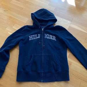 Marinblå hoodie från Tommy Hilfiger - Snygg marinblå hoodie från Tommy Hilfiger med broderad logga över bröstet. Tröjan har dragkedja, huva med snörning och två fickor framtill. Perfekt för chill dagar och enkel att matcha med jeans! Storleksmässigt så är den (S) enligt mig. Skicket är bra, en stilren tröja helt enkelt! Skriv om du har några funderingar/frågor. Mvh Oliver