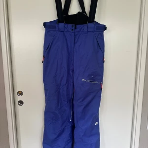 Blå täckbyxor från Everest, stl 40 - Blå täckbyxor från Everest med hängslen och flera dragkedjeförsedda fickor. Byxorna har justerbar midja med kardborre och är perfekta för kalla dagar. Snygg sportig look och praktiska detaljer för vinterns äventyr.