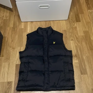 Svart dunväst från Lyle & Scott - Svart dunväst från Lyle & Scott med klassisk gul logga på bröstet. Västen har hög krage, två stora fickor framtill och quiltad design. Perfekt för lager-på-lager och enkel att matcha med olika outfits.