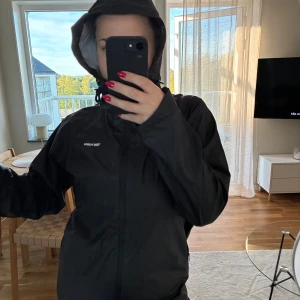 Svart regnjacka från Tretorn med huva - Svart regnjacka från Tretorn med huva och dragkedja framtill. Jackan är i ett slätt, vattentätt material och har diskret logga på bröstet. 