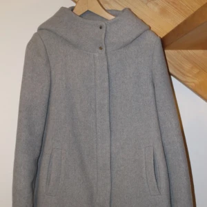 Grå ullkappa - Stilren grå kappa i ullblandning med mysig luva och dold dragkedja framtill. Kappan har två fickor och knappar vid halsen för extra detalj. Sååå mysig och perfekt till höst och vinter. M men passar xs/s om man vill ha lite oversized  