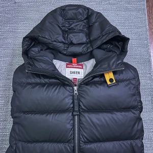 Svart pufferjacka Parajumpers Sheen - Svart pufferjacka från Parajumpers, modell Sheen. Jackan har huva, två dragkedjefickor och ikonisk gul detalj vid halsen. Broderad PJS-logga på ärmen. Tillverkad i glansig polyester och fodrad för extra värme. Perfekt för kalla dagar. Stl S 