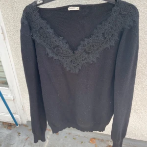 Svart stickad tröja med spets från Moocci - Svart stickad tröja från Moocci med djup v-ringning och bred spetsdetalj runt halsen. Långärmad och ribbad i materialet, perfekt för dig som gillar en klassisk men ändå unik look. Normal i storleken men lite oversized.
