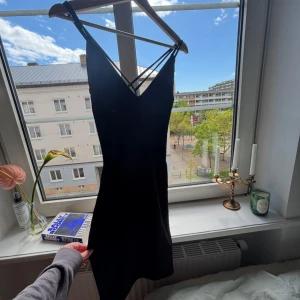 Svart kort klänning med smala axelband - Säljer en svart kort klänning med smala dubbla axelband som korsas i ryggen. Klänningen har v-ringning och figurnära passform, perfekt för dig som gillar minimalistisk och stilren look. Materialet känns mjukt och följsamt mot kroppen.