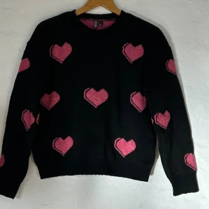 Svart stickad tröja med rosa hjärtan - Supersöt dubbelvaddrad stickad tröja med hjärtan på. Super varm, perfekt till kalla vinterdagar! Säljer då den inte kommer till användning längre. Lite nopprig men går lätt att få att se ny ut igen med en noppborttagare.