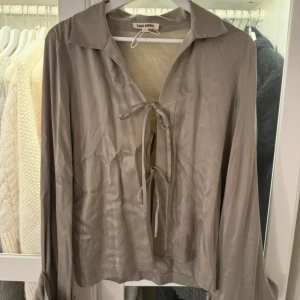  Djerf Avenue blus - Supersnygg beige blus från Djerf Avenue med långa ärmar och två knytningar framtill. Blusen har en klassisk krage och är gjord i ett mjukt, glansigt material som känns lyxigt. Perfekt att styla med jeans eller kjol för en trendig look.