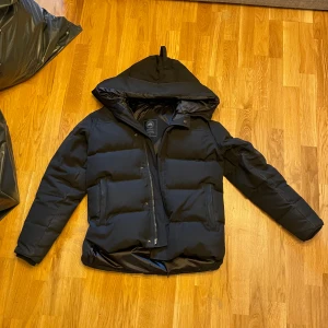 Canada Goose MacMillian vinterjacka - Säljer en svart vinterjacka från Canada Goose med huva och klassisk logga på ärmen. Jackan har dragkedja framtill, två stora fickor och quiltad design. Perfekt för kalla dagar och riktigt snygg streetstil.