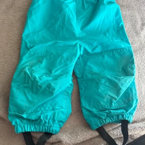 Turkosa täckbyxor från Kaxs stl 80 - Turkosa täckbyxor från Kaxs i storlek 80. Byxorna har resår i midjan och vid bensluten samt svarta fotremmar. Perfekta för kalla dagar och utomhuslek. Materialet är slitstarkt och vattenavvisande syntet.