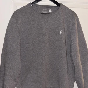 Grå sweatshirt från Polo Ralph Lauren - En stilren grå sweatshirt från Polo Ralph Lauren med klassisk rund halsringning och broderad vit logga på bröstet. Tröjan har långa ärmar och är tillverkad i mjuk bomullsmix, perfekt för en chill och avslappnad look. ( första två bilderna är med blixt )